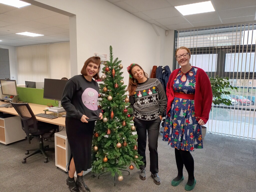 Three women in christmas jumpers stood by a christmas tree